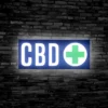 Illuminated CBD LED Sign with a green cross, hanging up on a black brick wall