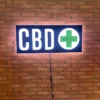 Lit up CBD LED sign with a green cross mounted on a red brick wall