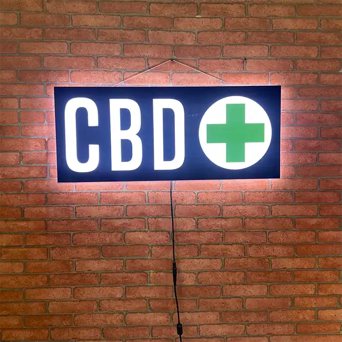 Lit up CBD LED sign with a green cross mounted on a red brick wall