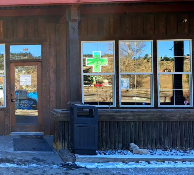 A storefront with a wooden exterior displays a green pharmacy cross and "open" sign in the window; a trash bin is placed by the entrance.